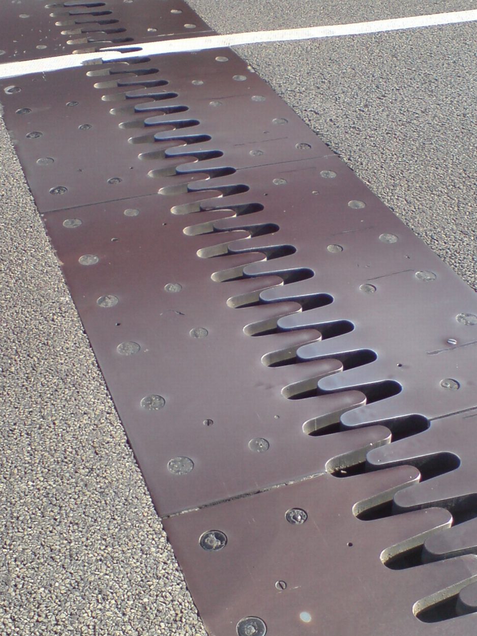 A close-up view of a metal bridge expansion joint with interlocking finger plates embedded in an asphalt road, featuring a white line in the background.