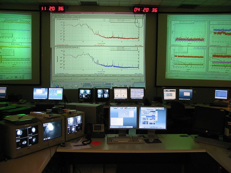 This figure shows a windowless room full of desks and computer screens and with three large screens on the wall upon which are projected a lot of technical graphs.