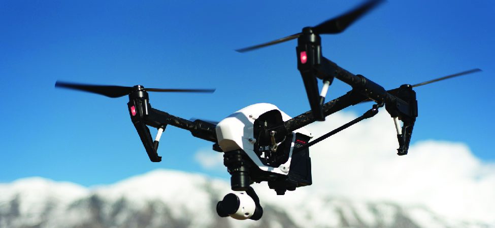 A white and black drone with its propellers spinning is captured in mid-flight against a clear blue sky, with a range of snow-capped mountains visible in the blurry background.