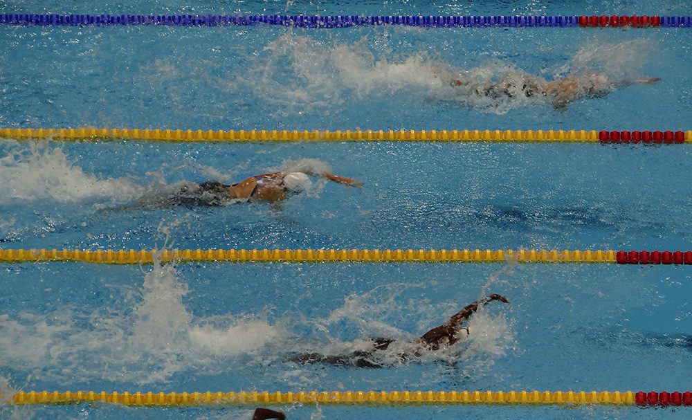 Swimmers are moving through separate pool lanes, with coloured lane ropes and splashing water.