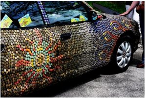 Photo of the side of a small four-door car whose bodywork is tiled edge to edge with bottle caps: front fender, both doors, rear quarter panel and the pillars between the windows are all covered, leaving only the glass, wheels and tyres bare. The caps are mostly gold, with paler ones laid out as a sunburst about a dozen caps across on the front door.