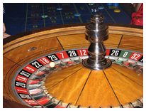Photograph of a roulette wheel seen at an angle from above, with the polished centre spindle in the middle and part of the numbered rim in view. The visible pockets alternate red and black and read 22, 18, 29, 7, 28, 12 and 26, followed by a green 0 and then 32, with the white ball sitting on the track and the blue betting layout out of focus behind.