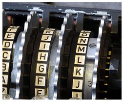 Close-up photograph of three alphabet rings inside an Enigma machine, standing side by side with notched metal ratchet wheels between them and a small numeral 3 stamped beside the right hand rotor. Only a short arc of each ring is in view, reading upward as A, B, C, D, E on the left ring, F, G, H, I, J, K on the middle ring and J, K, L, M, N, O on the right; the remainder of each alphabet is hidden inside the machine and cannot be read from this image.