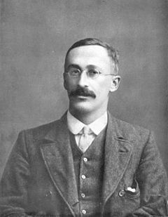 Black-and-white portrait photograph of William Sealy Gosset, a man with round wire glasses and a mustache, wearing a three-piece tweed suit and tie.